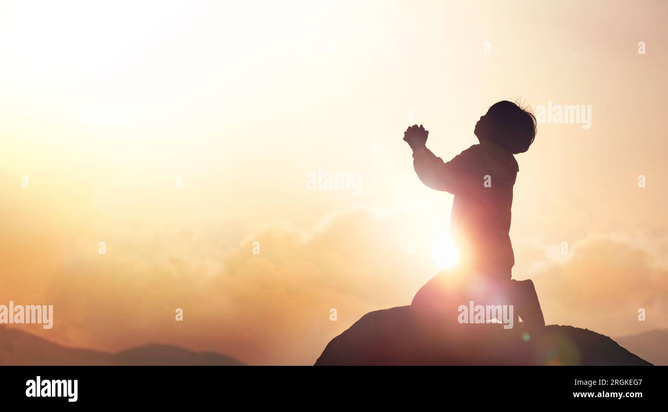 Child Christian earnestly praying to God and Jesus with both hands together under the red sunset sky and bright sunlight Stock Photo