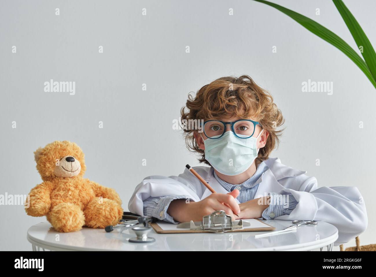 Child in eyeglasses and respiratory mask looking at camera while sitting at table with soft bear and clipboard on white background Stock Photo