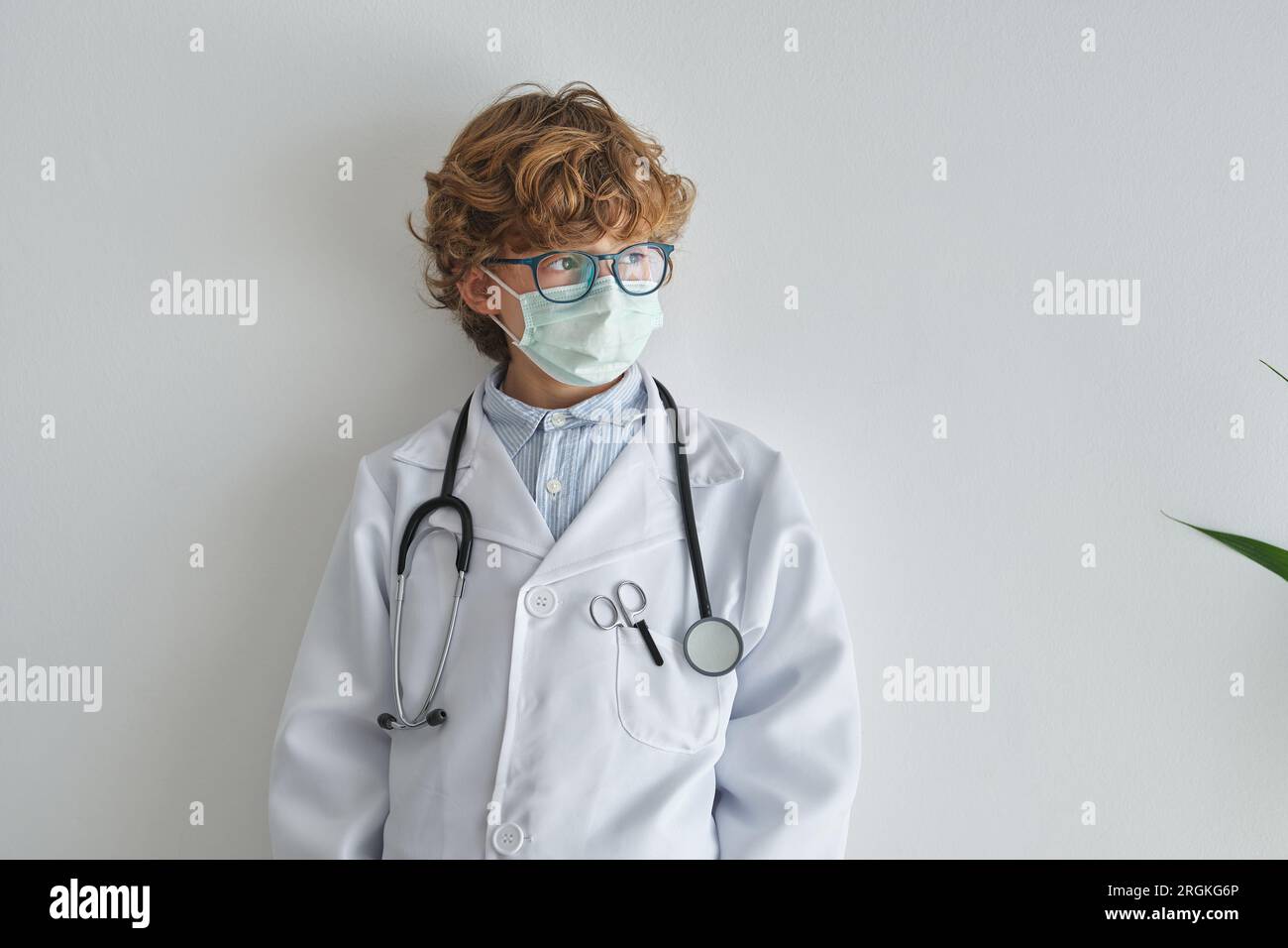 Contemplative child in eyeglasses and disposable mask with stethoscope on medical uniform looking away on white background Stock Photo