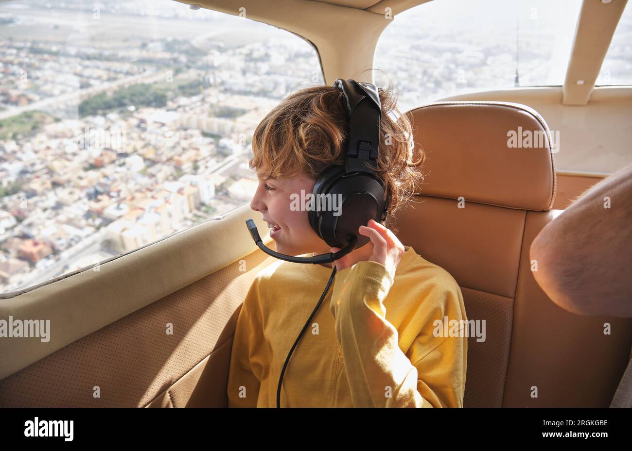 Cheerful boy with curly blond hair in casual clothes and headset smiling while sitting on passenger seat of modern small aircraft and admiring citysca Stock Photo