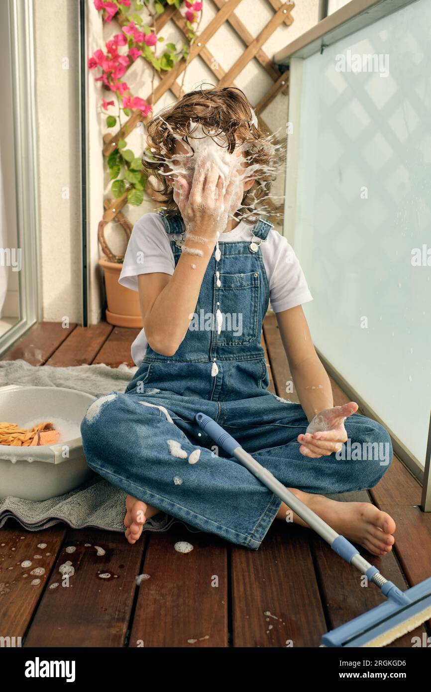 Barefoot anonymous preteen boy sitting on wooden balcony floor and hitting face with foam splashes while washing glass in sunlight Stock Photo