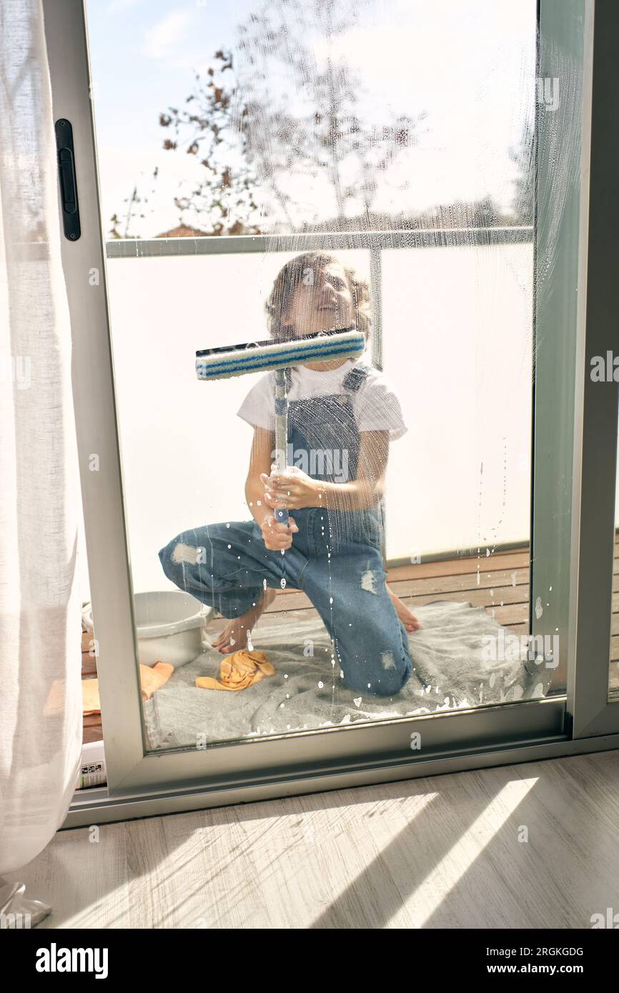 Full body of child in denim overall sitting on towel and looking up while doing household chores and wiping foam off glass door on balcony at home Stock Photo