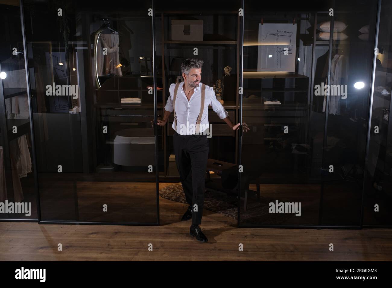 Middle aged bearded man in white shirt and black trousers with suspenders standing in doorway of dressing room Stock Photo