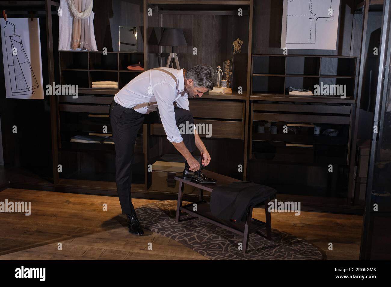 Side view of full length of gray haired man in elegant clothes tying laces on wooden stool and looking down while spending time in atelier Stock Photo
