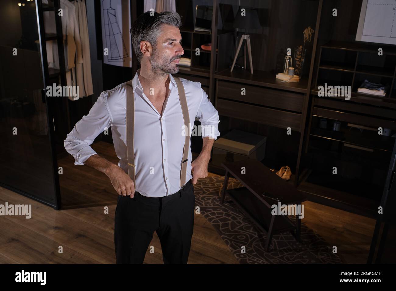 High angle of self assured unshaven Hispanic man in classy outfit looking away and standing with hands on waist against wooden shelves while having ti Stock Photo