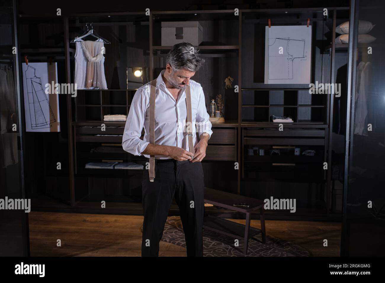 Serious middle aged Hispanic man in elegant clothes looking down and putting on suspenders while standing near wooden shelves in atelier Stock Photo