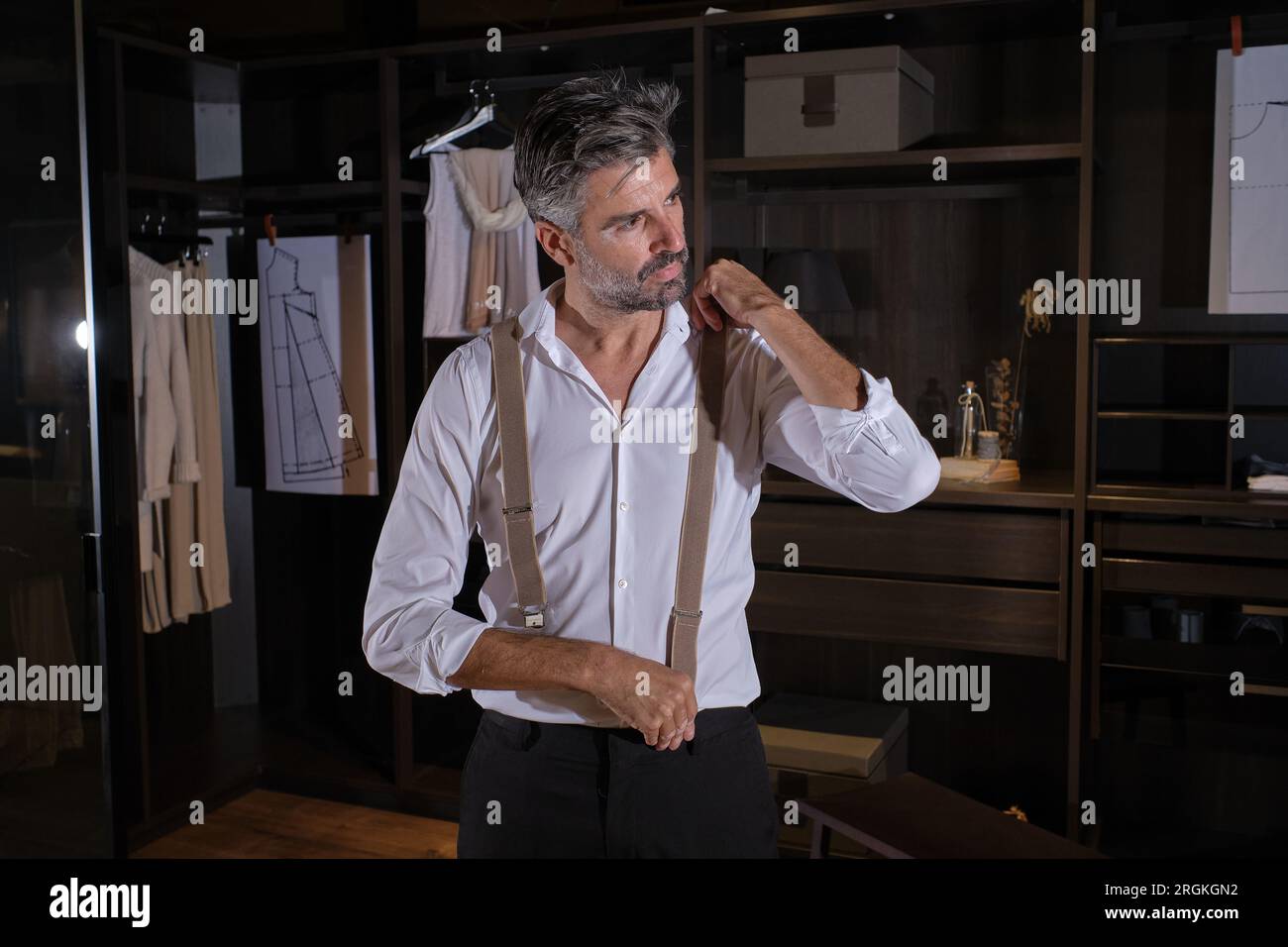 Serious unshaven Hispanic man wearing white shirt black trousers and suspenders looking away while standing in dark dressing room Stock Photo