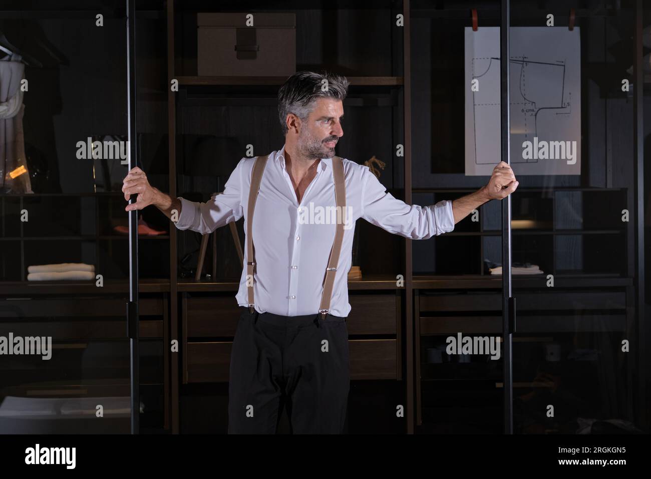 Mature man in white shirt and trousers with suspenders looking away while standing near wardrobe in dim light Stock Photo