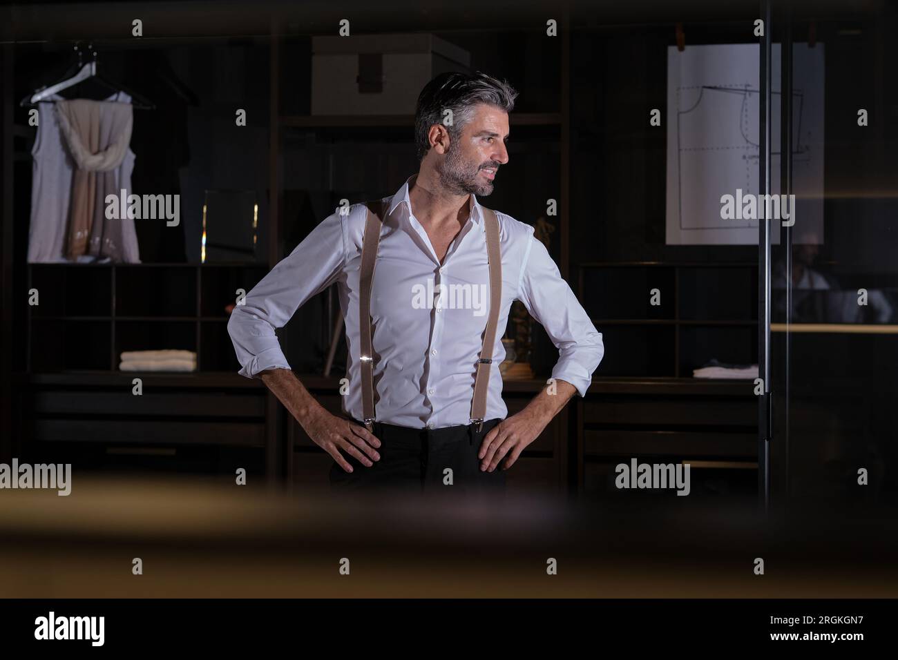 Glad middle aged man in white shirt and black pants with suspenders looking away with hands on waist at home Stock Photo