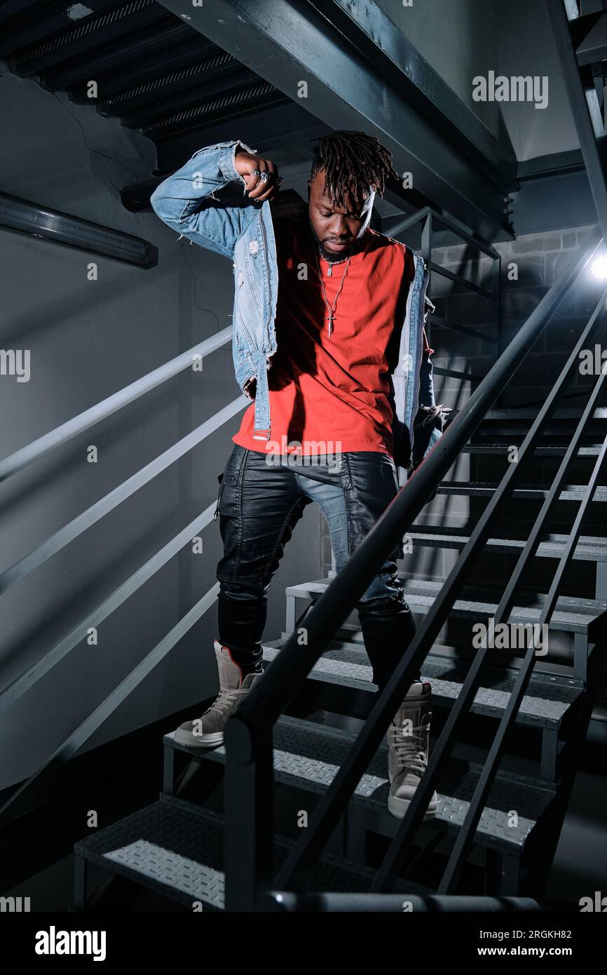 Young stylish black male looking down and putting on jeans jacket while standing on steps in dim light Stock Photo