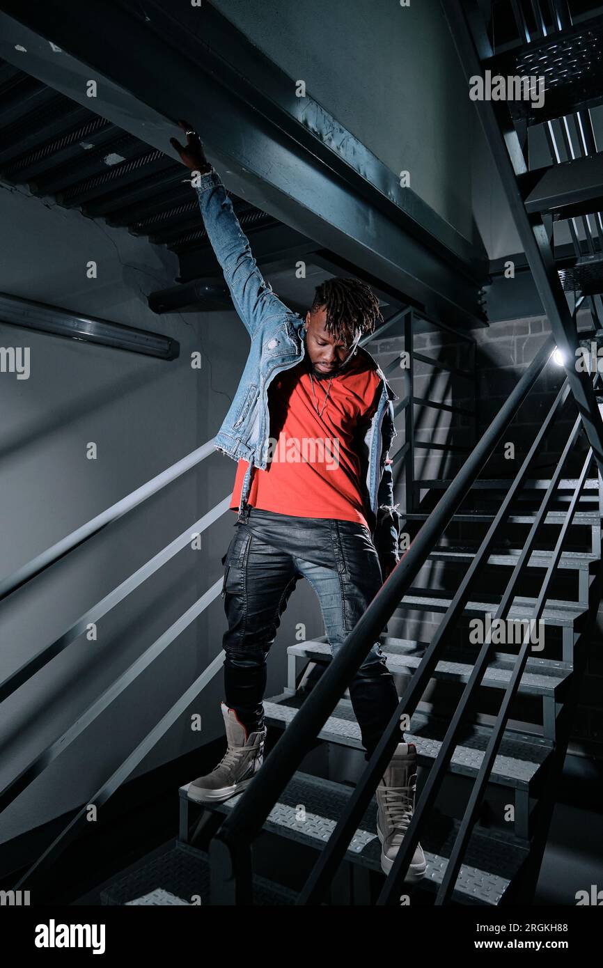 Full body of black man standing on metal stairs in dark building and looking down while putting on denim jacket Stock Photo