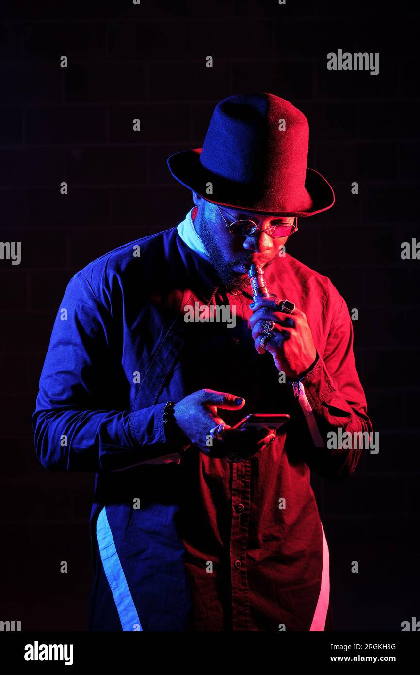 Black male model in trendy outfit with hat smoking e cigarette and chatting via phone on dark background in neon lighting Stock Photo