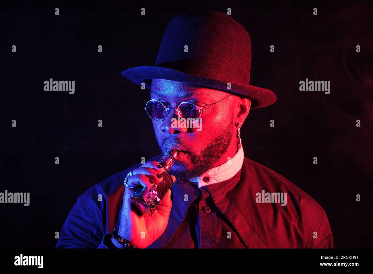 Confident African American male model in trendy black attire smoking e cigarette while standing on dark background in neon lighting Stock Photo