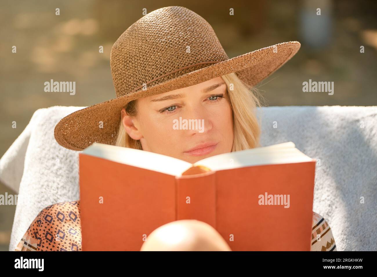 Pensive young female in straw hat and jacket looking at red bounded book while reading with concentration and sitting on white mattress on deckchair Stock Photo