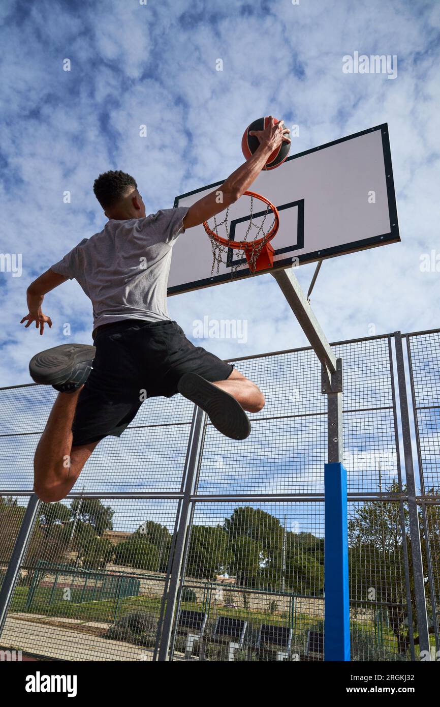 Low angle back view of African American male basketball player jumping above ground and throwing ball while playing basketball on sports ground Stock Photo