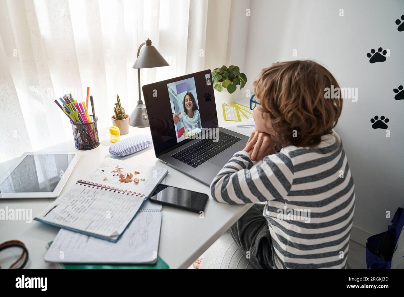 Side view of preteen boy with curly hair sitting at table and having video chat with friend via netbook while doing homework in room Stock Photo