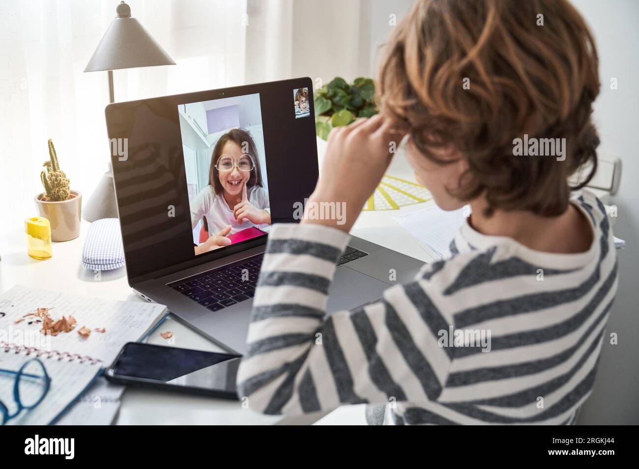 Side view of kid video chatting on laptop with girl in eyeglasses while sitting at table with smartphone in room Stock Photo