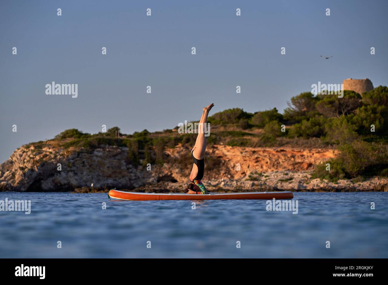 Side view of sporty female athlete in swimsuit taking Salamba Shirshasana A pose on SUP board while practicing yoga among rippling blue ocean near roc Stock Photo