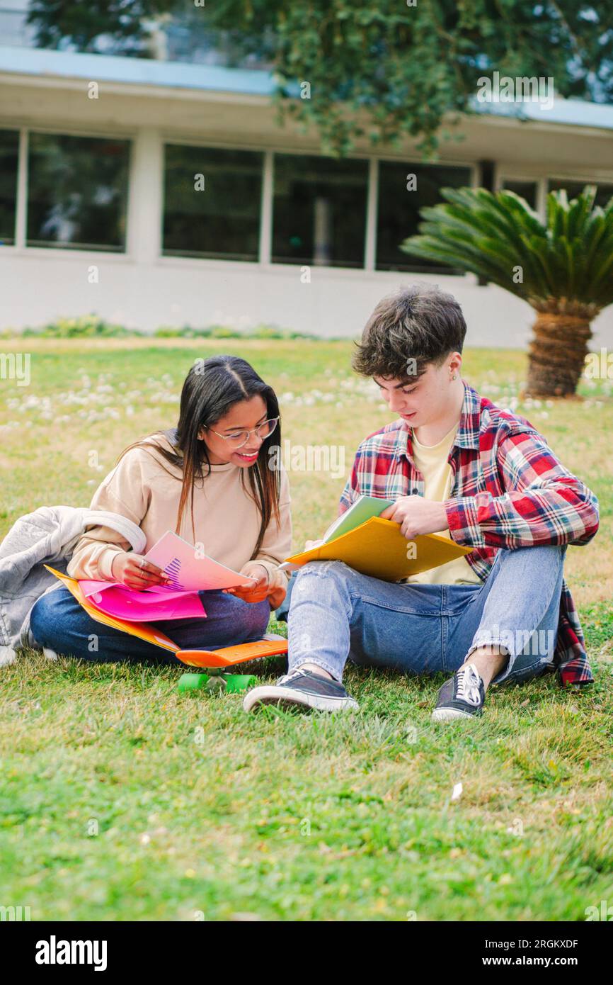 Vertical portrait of a student couple learning the lesson sitting on the lawn at high school university campus. A pair of young teenage friends doing the homeworks of the college class together. High quality photo Stock Photo