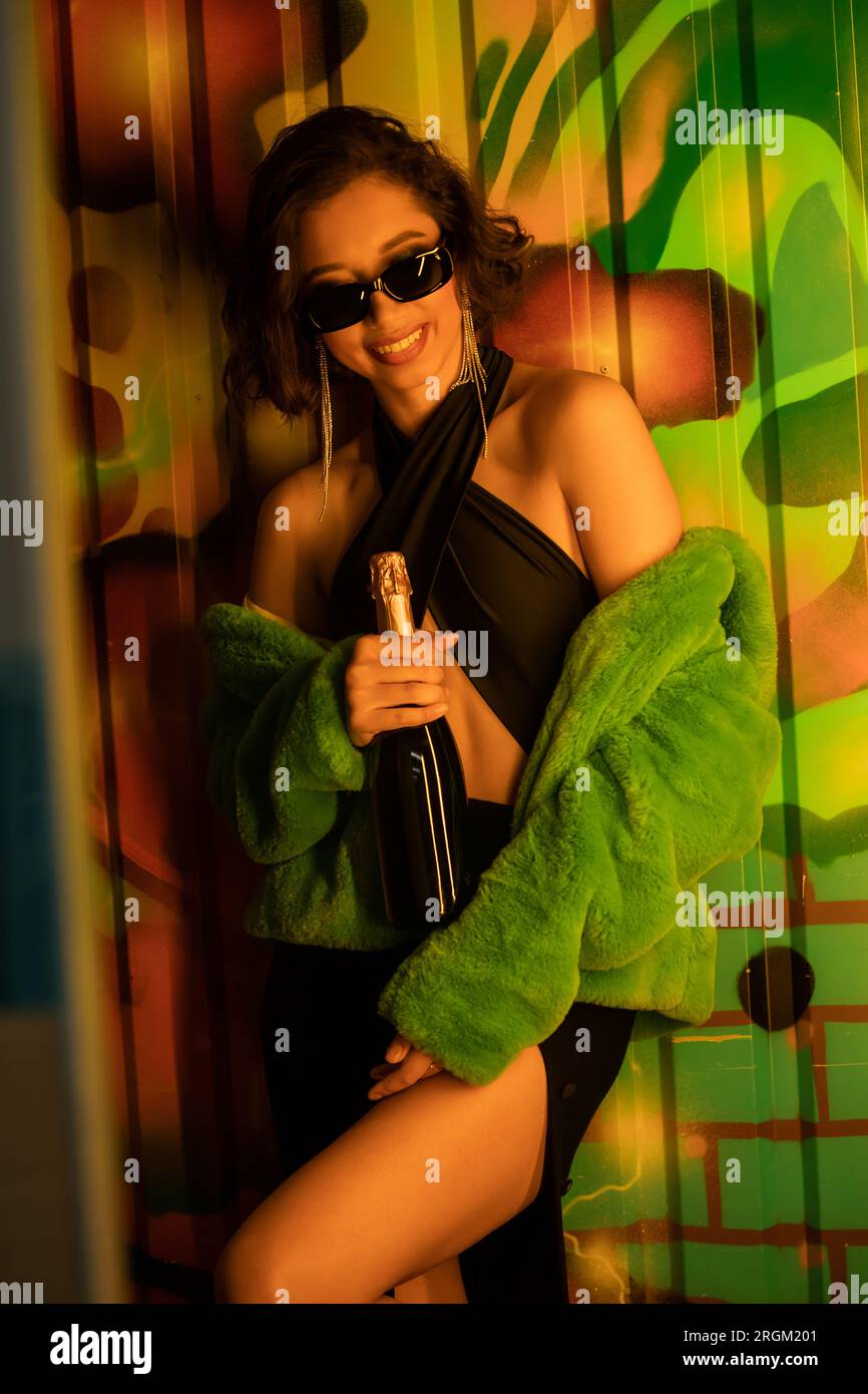 Cheerful asian woman in sunglasses and fake fur jacket holding champagne near graffiti in night club Stock Photo