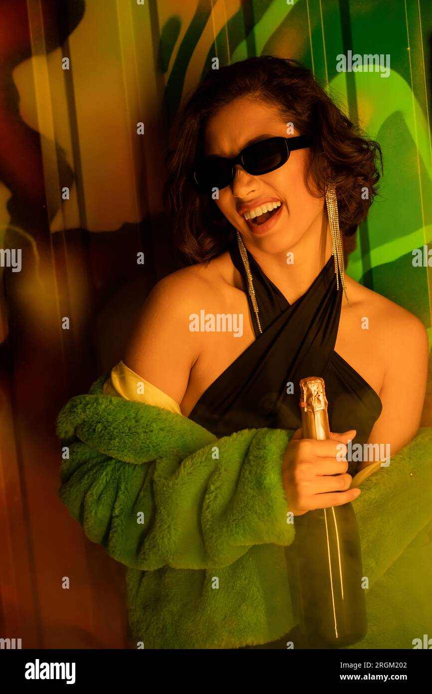 Excited asian woman in sunglasses and fake fur jacket holding champagne near graffiti in night club Stock Photo