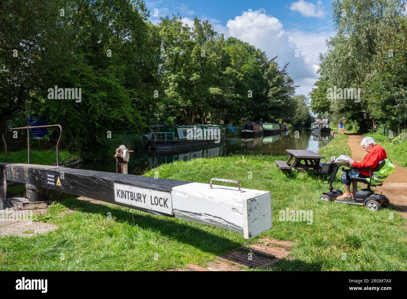 Kintbury Lock on the Kennet and Avon Canal in Berkshire, England, UK, with an old lady senior woman sitting on a mobility scooter reading a newspaper Stock Photo