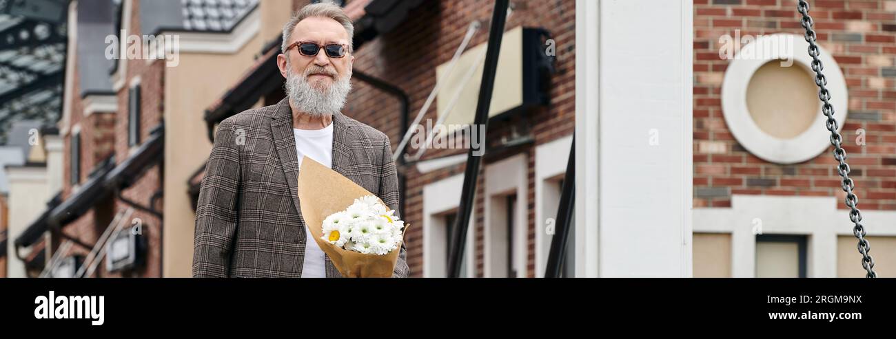 senior man with beard and sunglasses holding bouquet of flowers, standing on urban street, banner Stock Photo