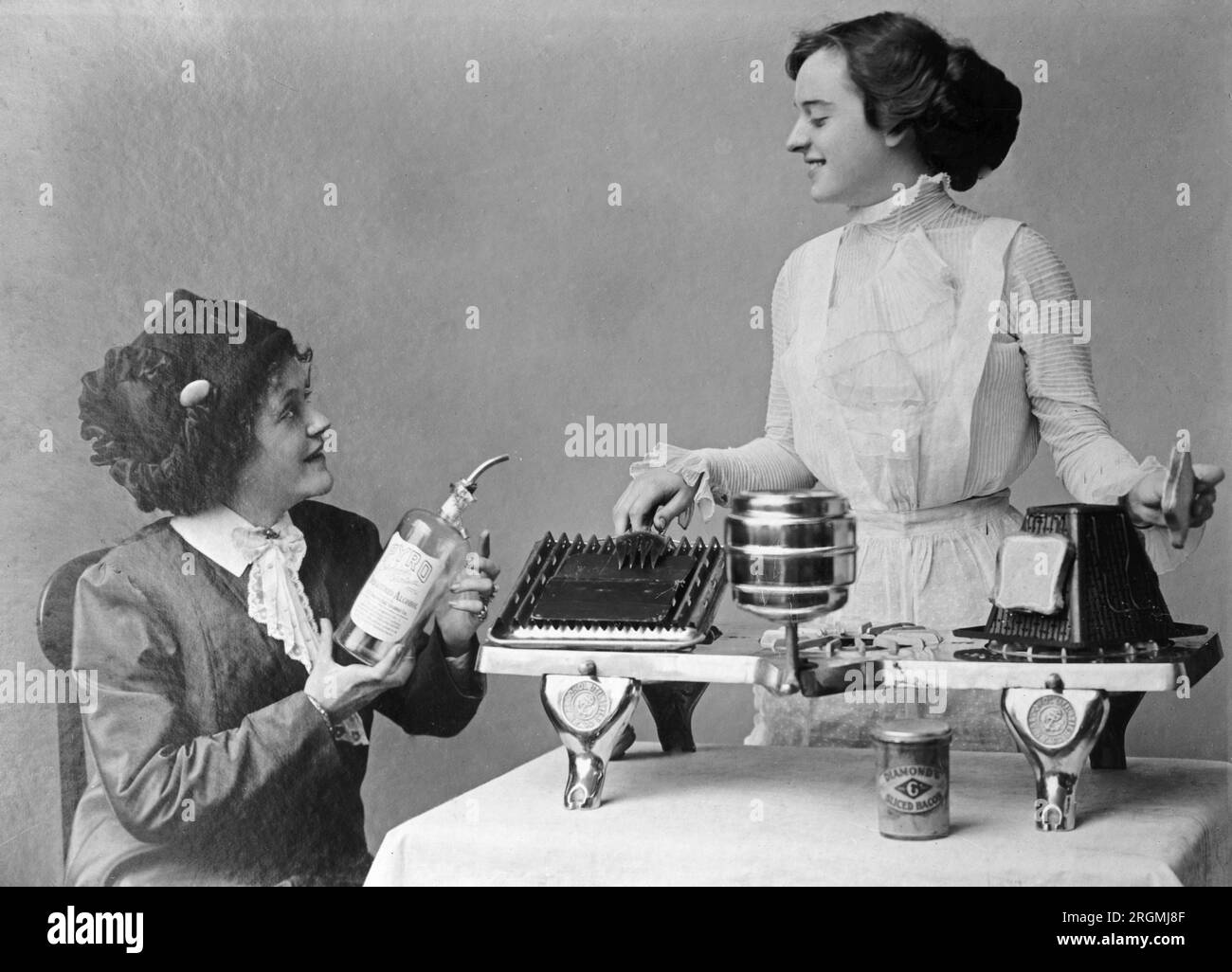 Photo shows a seated woman in a dress and hat holding a glass bottle with a pouring spout labeled 'Pyro' denatured alcohol. Another woman in a dress and apron stands behind a 3-burner appliance that can heat toast and other food ca. 1912-1930 Stock Photo