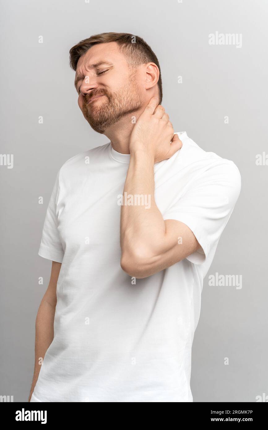 Unhappy frown man feels severe pain in neck trying to stretch muscles in spine on grey studio wall  Stock Photo