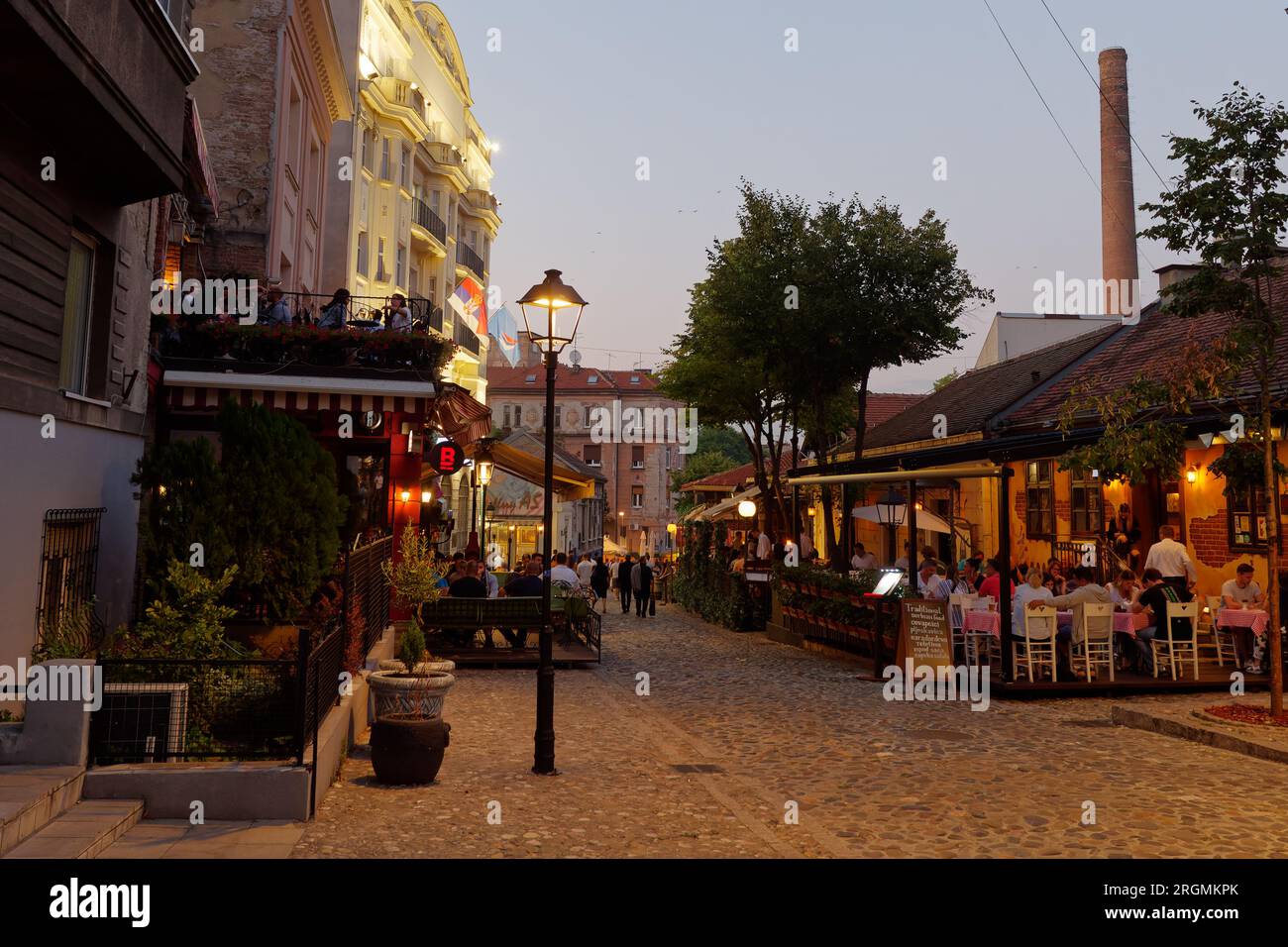 Bohemian cobbled Skadarlija street with people in cafes and restaurants. Chimney of old brewery right. Belgrade, Serbia. August 10, 2023.. Stock Photo