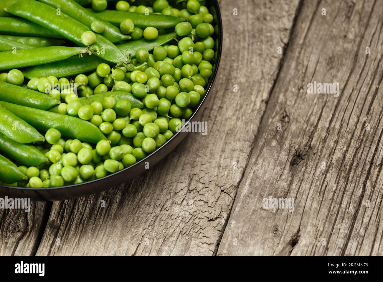 Green pea pods and shelled organic fresh peas in a round black plate on an aged wooden background, copy space. Vegetable protein, healthy products. Stock Photo