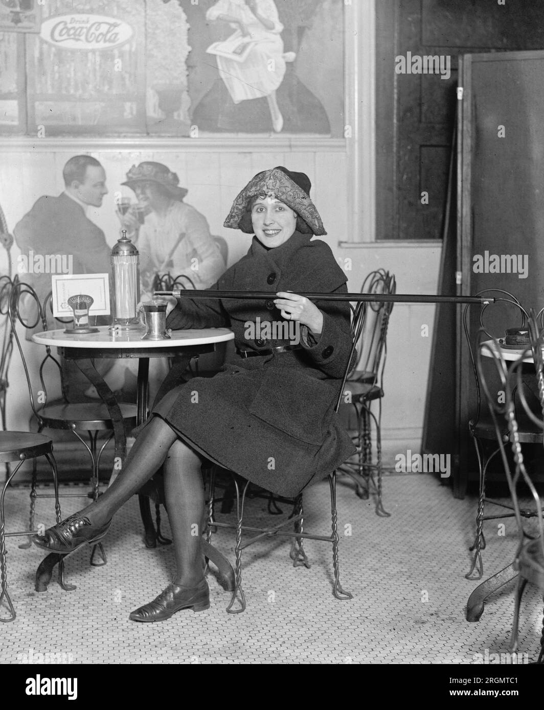 Woman seated at a soda fountain table is pouring alcohol into a cup from a cane, during Prohibition; with a large Coca-Cola advertisement on the wall ca. 1922 Stock Photo