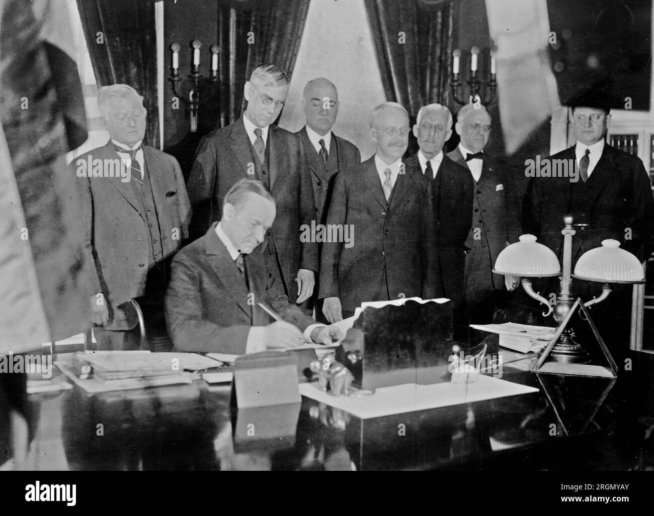 Calvin Coolidge signing the income tax bill, also known as the Mellon tax bill. Secretary of the Treasury Andrew Mellon is the third figure from the right, and Director of the Budget, General Herbert Mayhew Lord, is to Mellon's left ca. 1926 Stock Photo