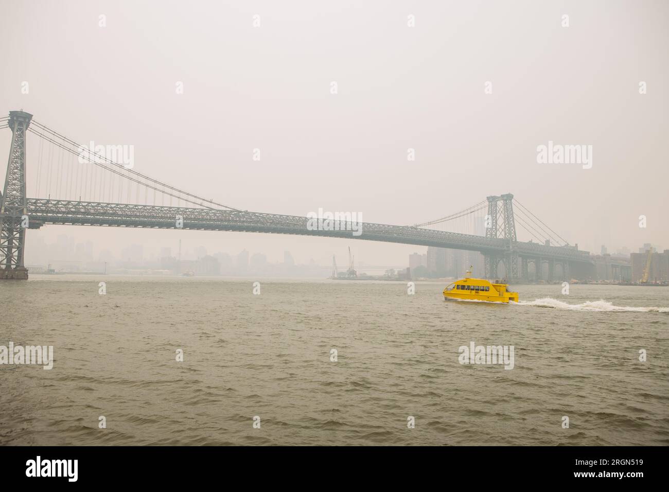 Williamsburg Bridge taken from the Domino Park, Williamsburg, Brooklyn, in a misty and foggy day. Yellow taxi boat passing under it. Stock Photo