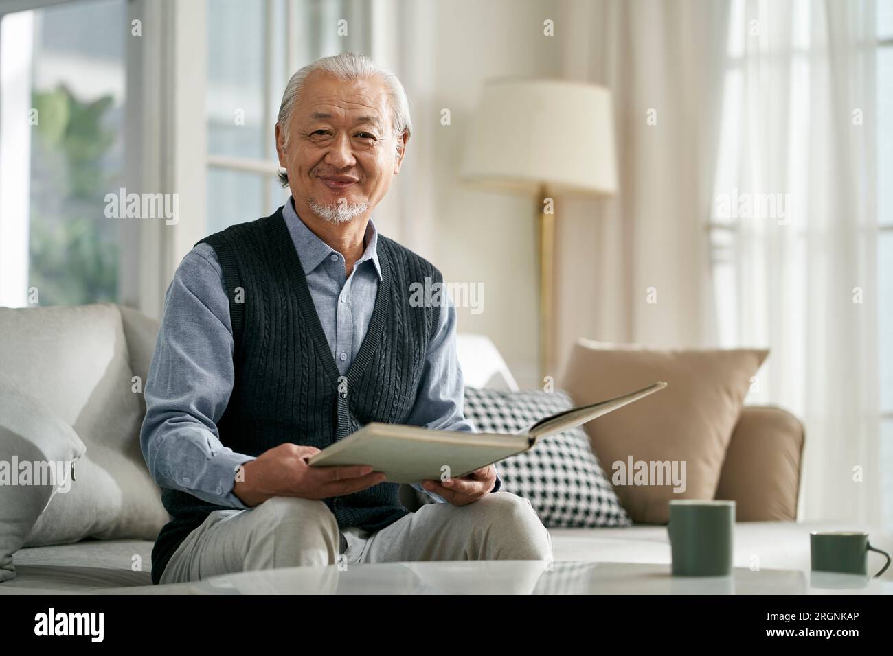 happy asian old man sitting on couch at home reading a book looking at camera smiling Stock Photo