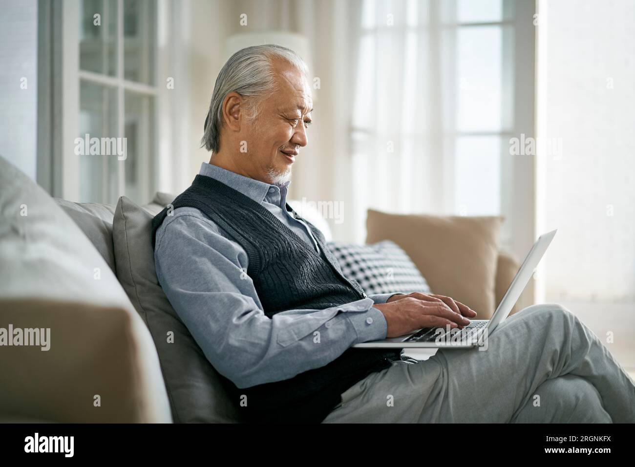 side view of senior asian man sitting on couch at home using laptop computer Stock Photo