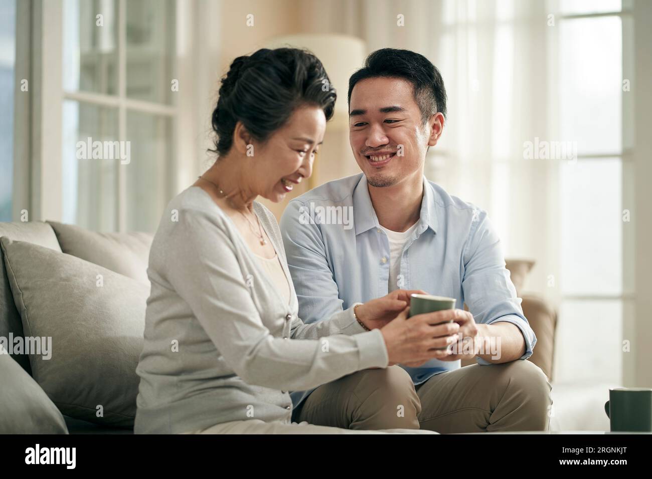asian adult son sitting on couch at home chatting with senior mother happy and smiling Stock Photo