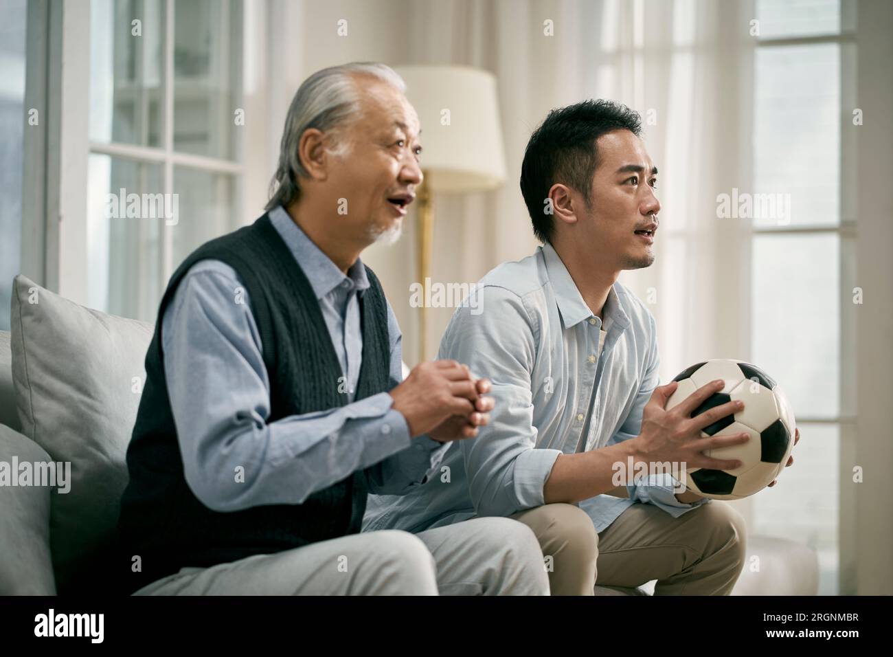 senior asian father and adult son sitting on couch at home watching soccer game on tv together Stock Photo