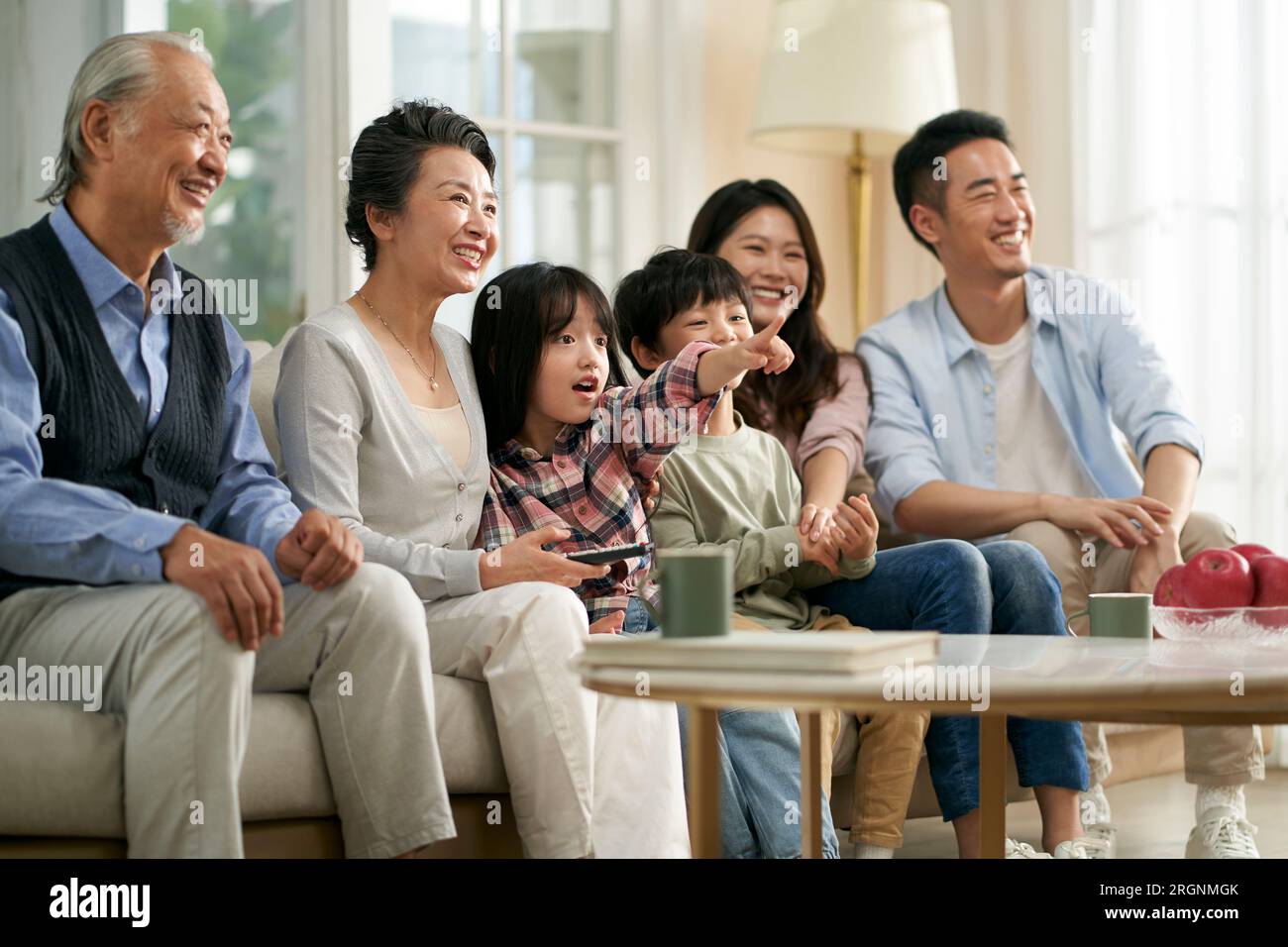 three generation asian family sitting on couch at home watching tv together happy and smiling Stock Photo