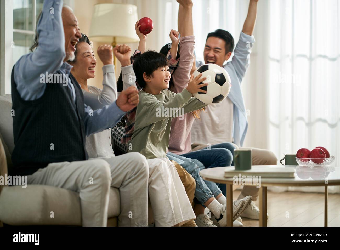 three generation asian family sitting on couch at home celebrating goal and victory while watching live broadcasting of football match Stock Photo