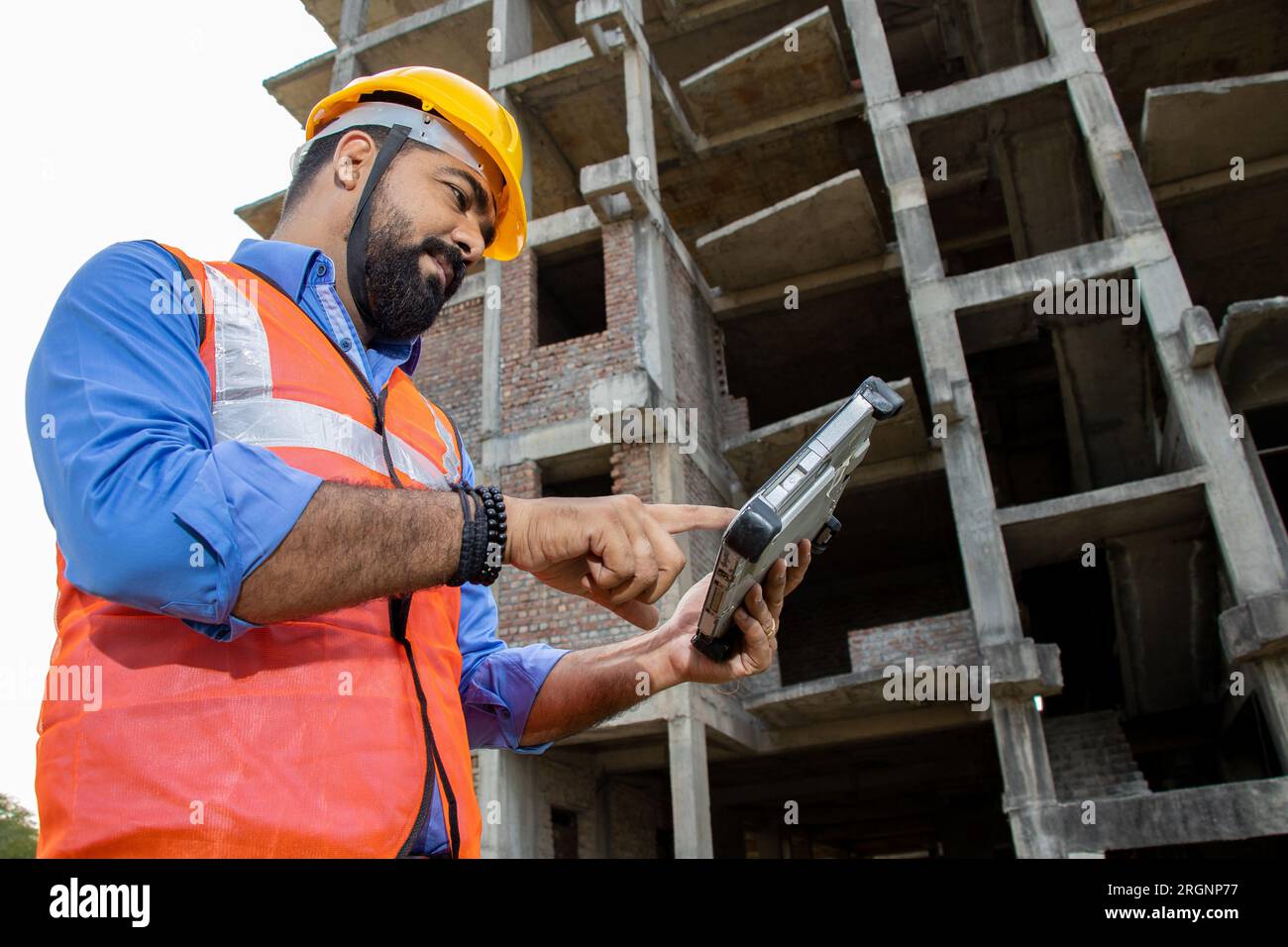 Young Indian male civil engineer or architect wearing helmet and vest holding digital tablet blueprint at construction site. Low angle. Stock Photo