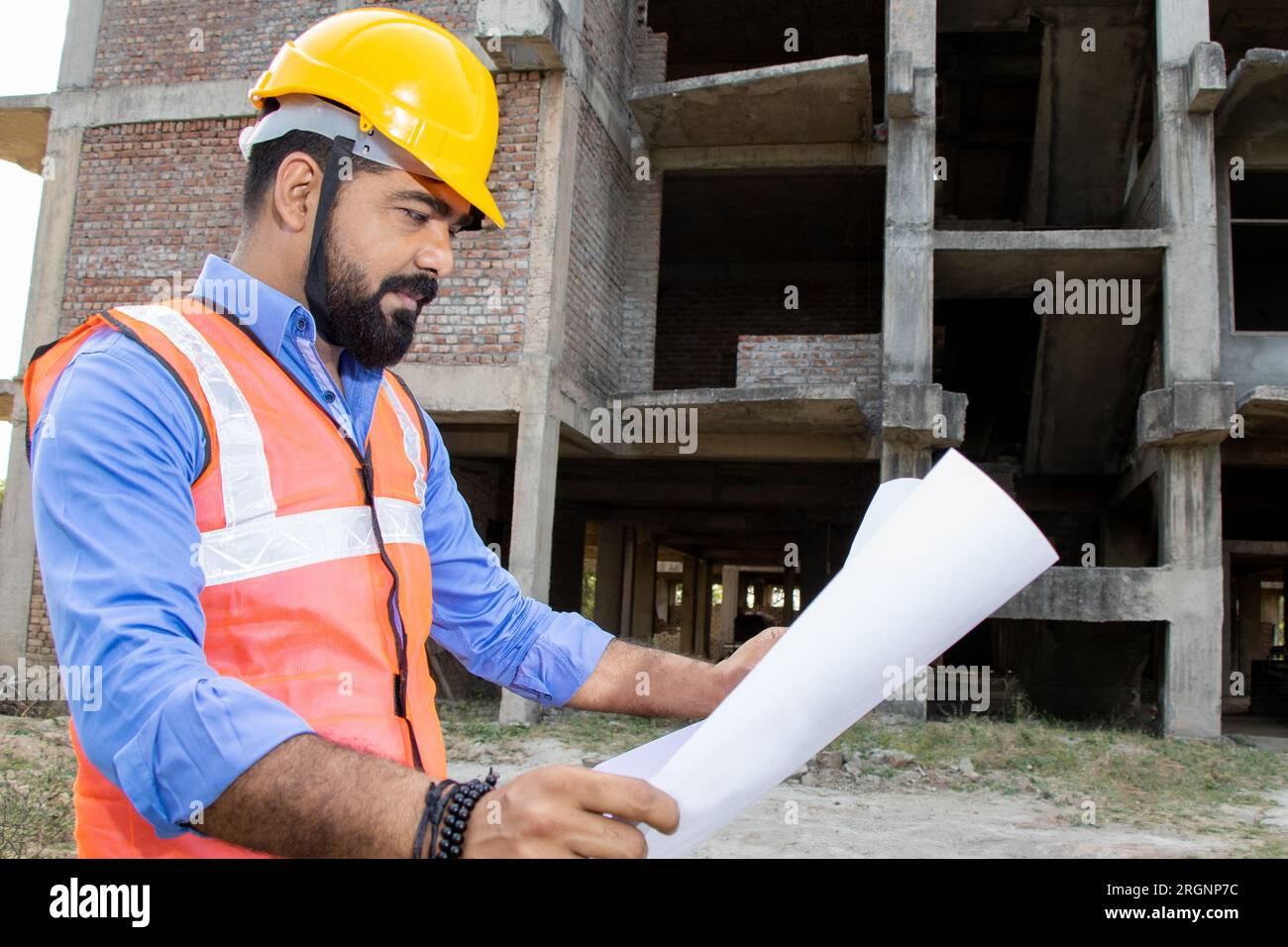 Young Indian male civil engineer or architect wearing helmet and vest holding paperwork blueprint at construction site. Stock Photo