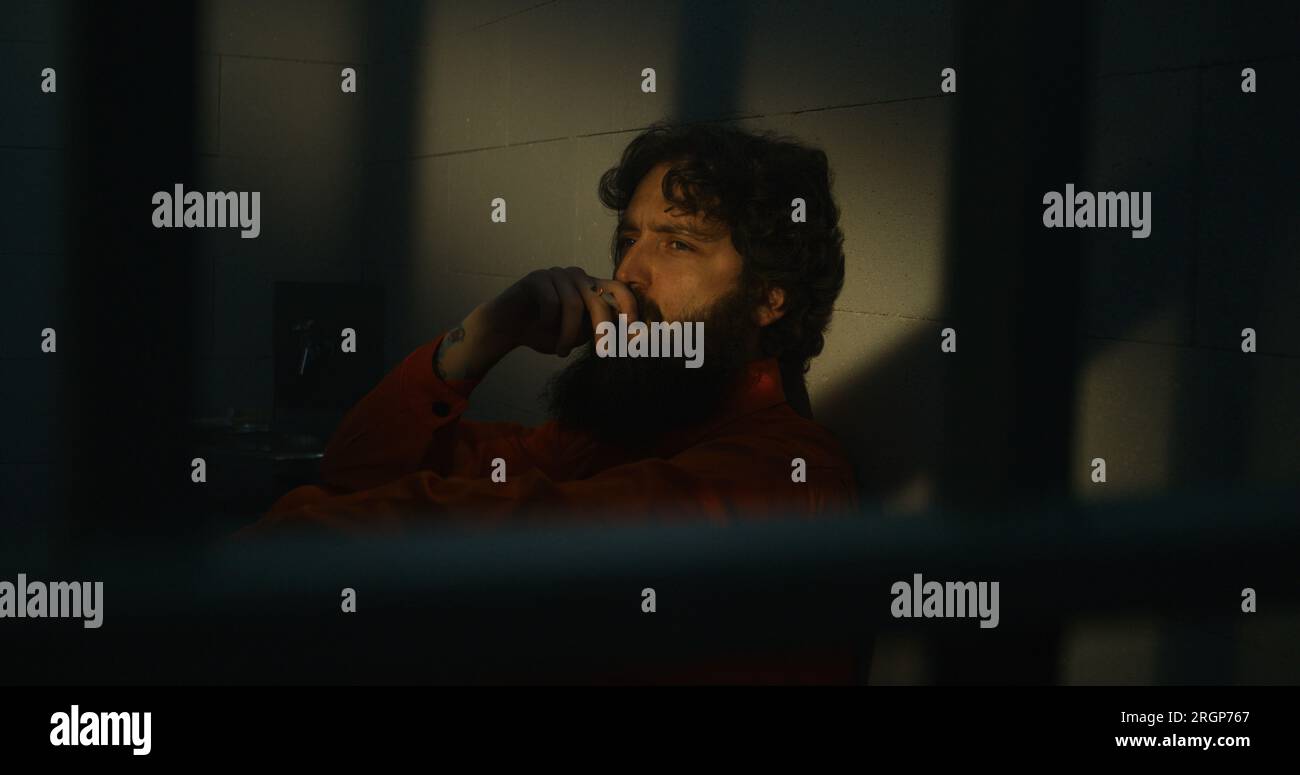 Depressed prisoner in orange uniform sits on bed, smokes cigarette in