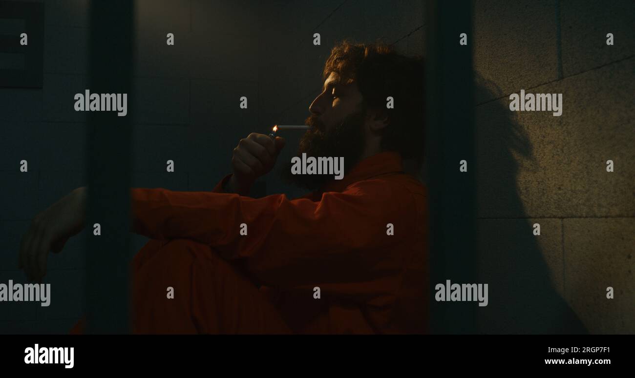 Depressed male prisoner in orange uniform sits on the bed, smokes