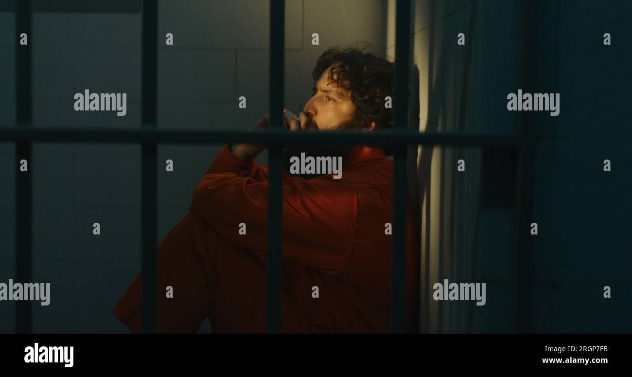 Pensive male prisoner in orange uniform sits on bed, smokes cigarette