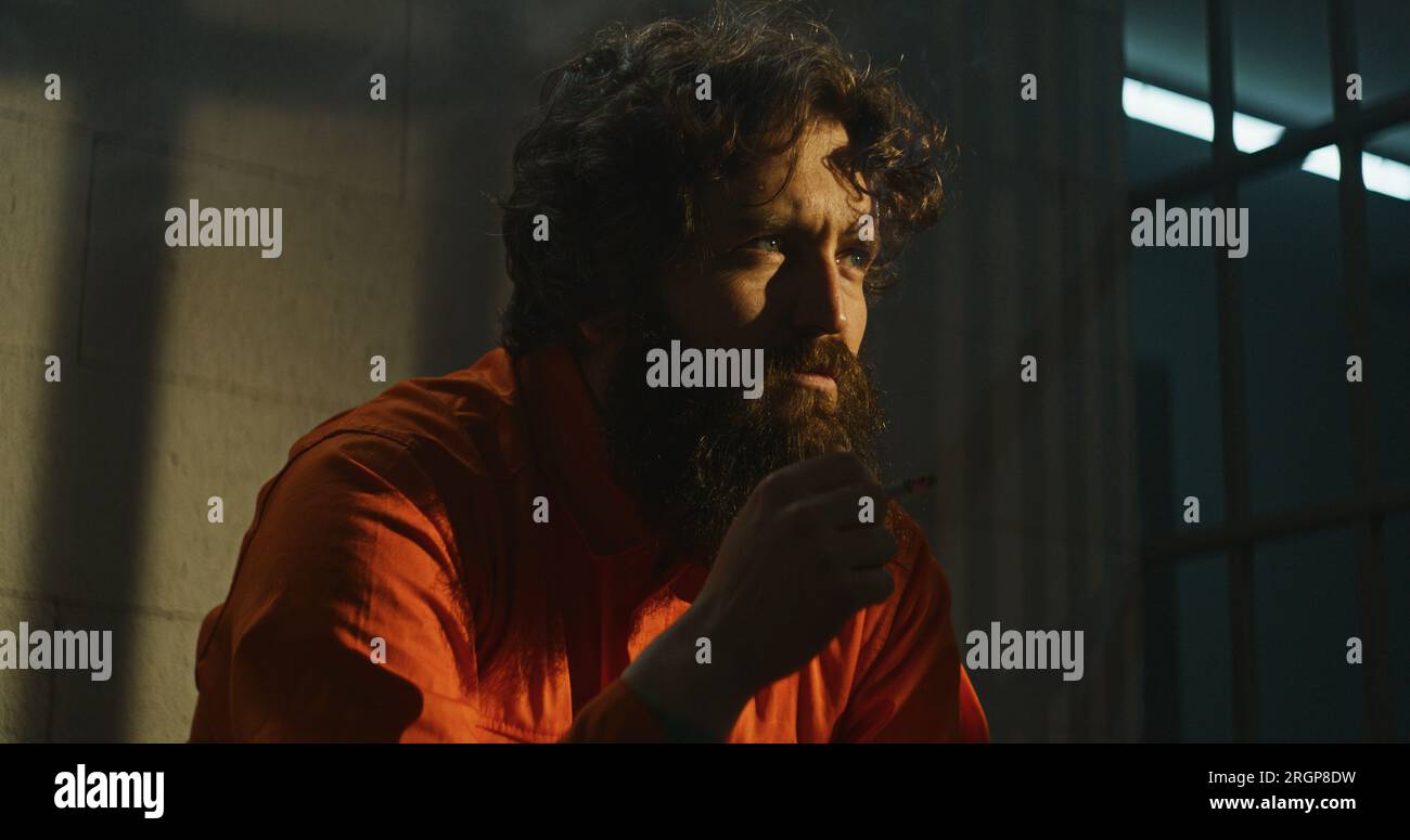 Depressed prisoner in orange uniform sits on bed in prison cell, smokes ...