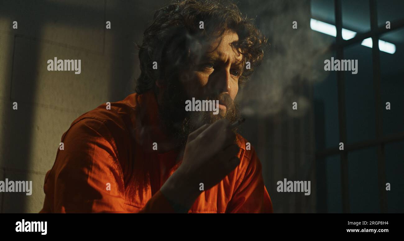 Depressed prisoner in orange uniform sits on bed in prison cell, smokes