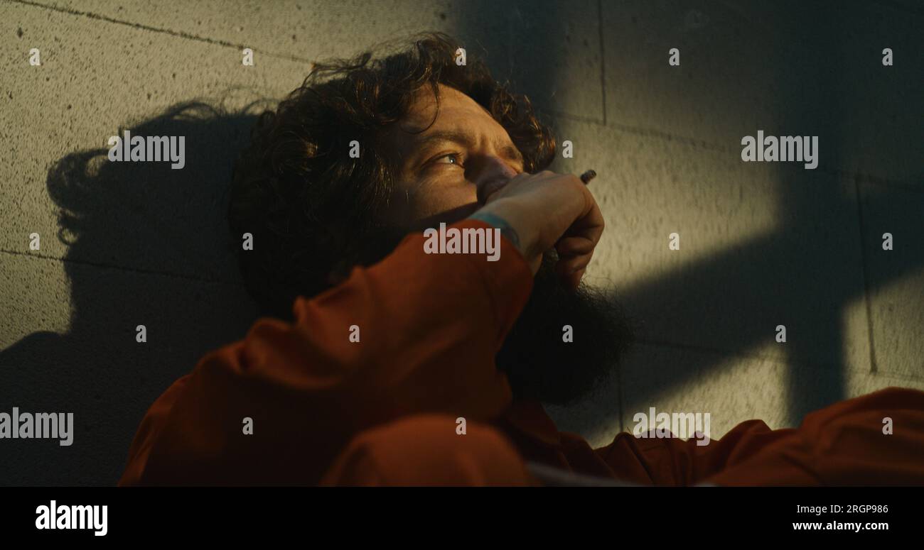 Depressed prisoner in orange uniform sits on bed in prison cell, smokes