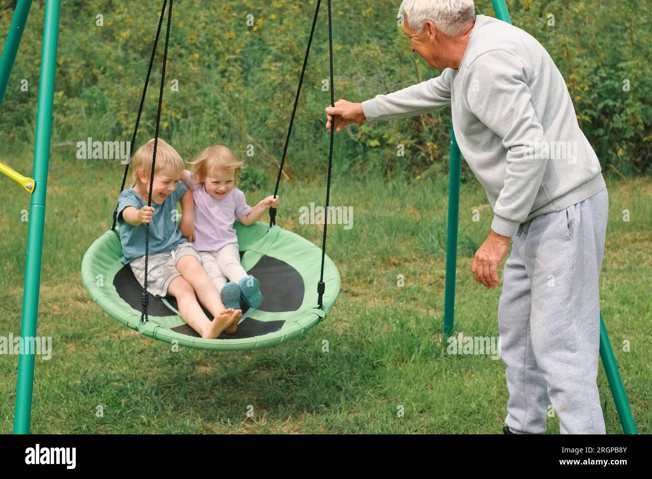 Grandfather swinging children in summer park. Grand dad and grandchildren sitting on swing outdoors. Senior 60s Grandpa pushing small grandkids on a r Stock Photo