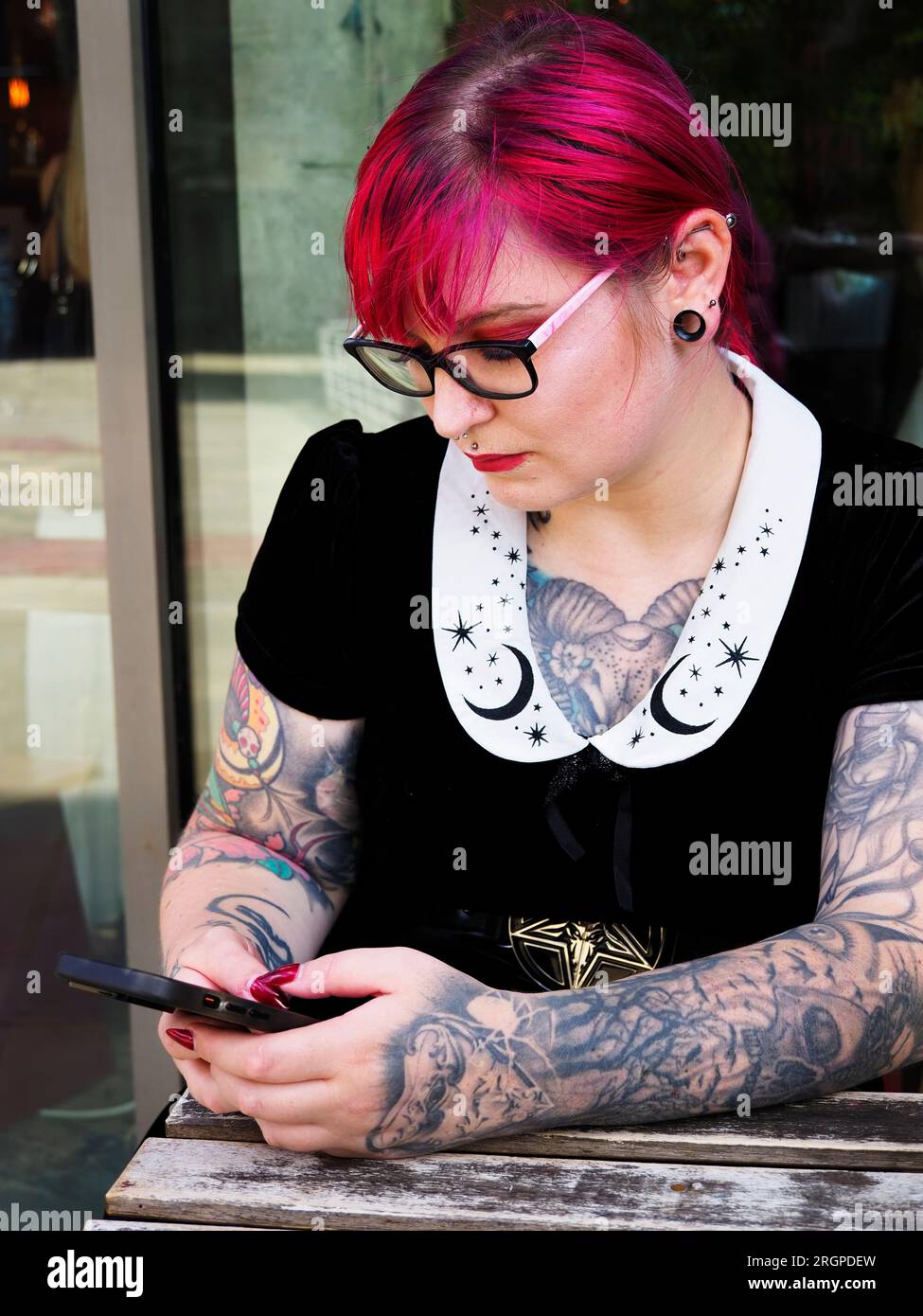 Woman in her mid twenties with dyed red hair and tattoos wearing glasses using a smartphone in Leeds West Yorkshire England Stock Photo